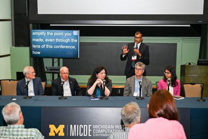 The panel sits at a long table, in front of which is a banner with the MICDE logo. Duraisamy stands, speaking.