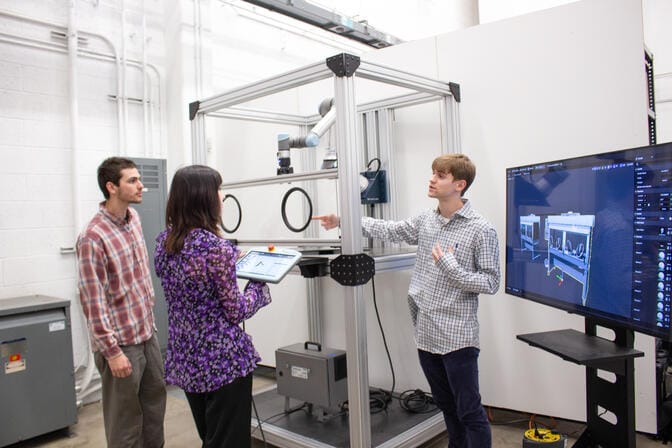 Three individuals stand around a robotic system frame in a lab while viewing and discussing its digital twin displayed on a nearby screen. One person holds a tablet, and another gestures while explaining the system.