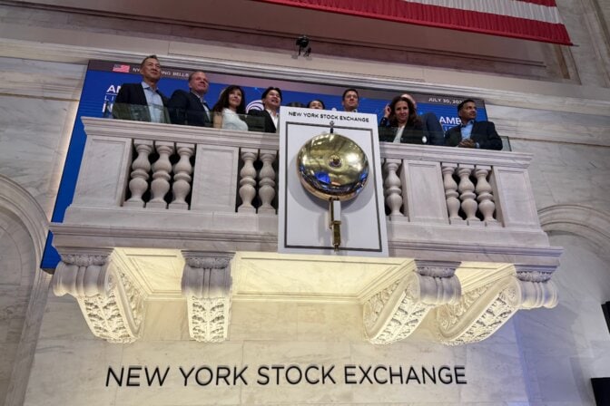 Men and women dressed in suits stand on a balcony behind a large gold bell. Below the balcony, lettering on the wall says New York Stock Exchange.