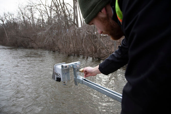 A person adjusts a device on a metal railing above a river with leafless trees in the background.