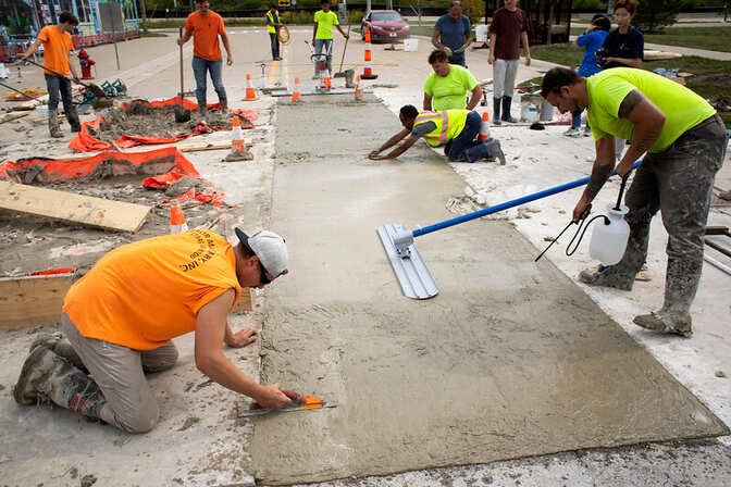 Construction workers smoothing wet concrete on a sidewalk with tools and safety cones around.