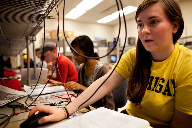 Students working with computers and electronics in a classroom, laboratory setting.The room is bright, with fluorescent lighting and shelves containing various equipment.
