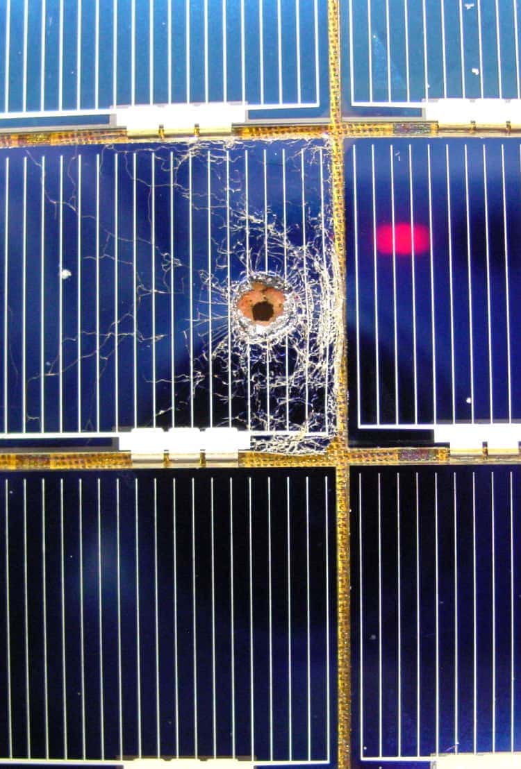 Six deep-blue solar panels are arranged in a grid that is three panels tall and two panels wide. A crater in the center-left panel marks the impact of a collision. Cracks stretch across the panel's glass-like surface, radiating out from the crater almost like a spider web.