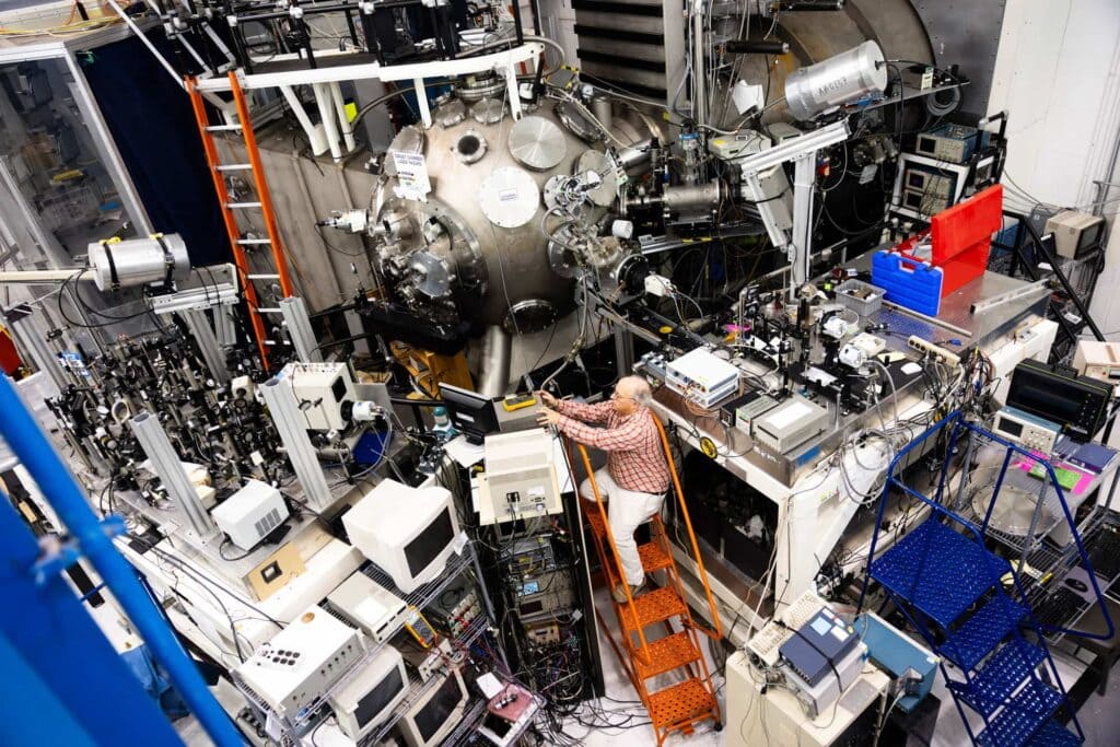 A man climbs an orange step ladder nestled among a tangle of electrical consoles, computer monitors, lenses, and wires. The extra height helps him reach a button on one console at the top of a stack of shelves. The entire collection of instruments border a large, metal orb in the center of the room.