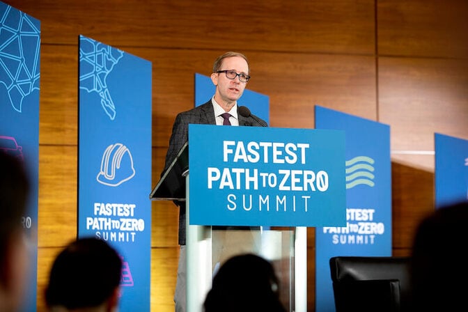 A speaker at a podium with a sign reading "FASTEST PATH TO ZERO SUMMIT," with banners in the background.