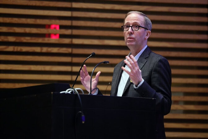 Todd Allen speaking at a podium with wooden slats in the background.