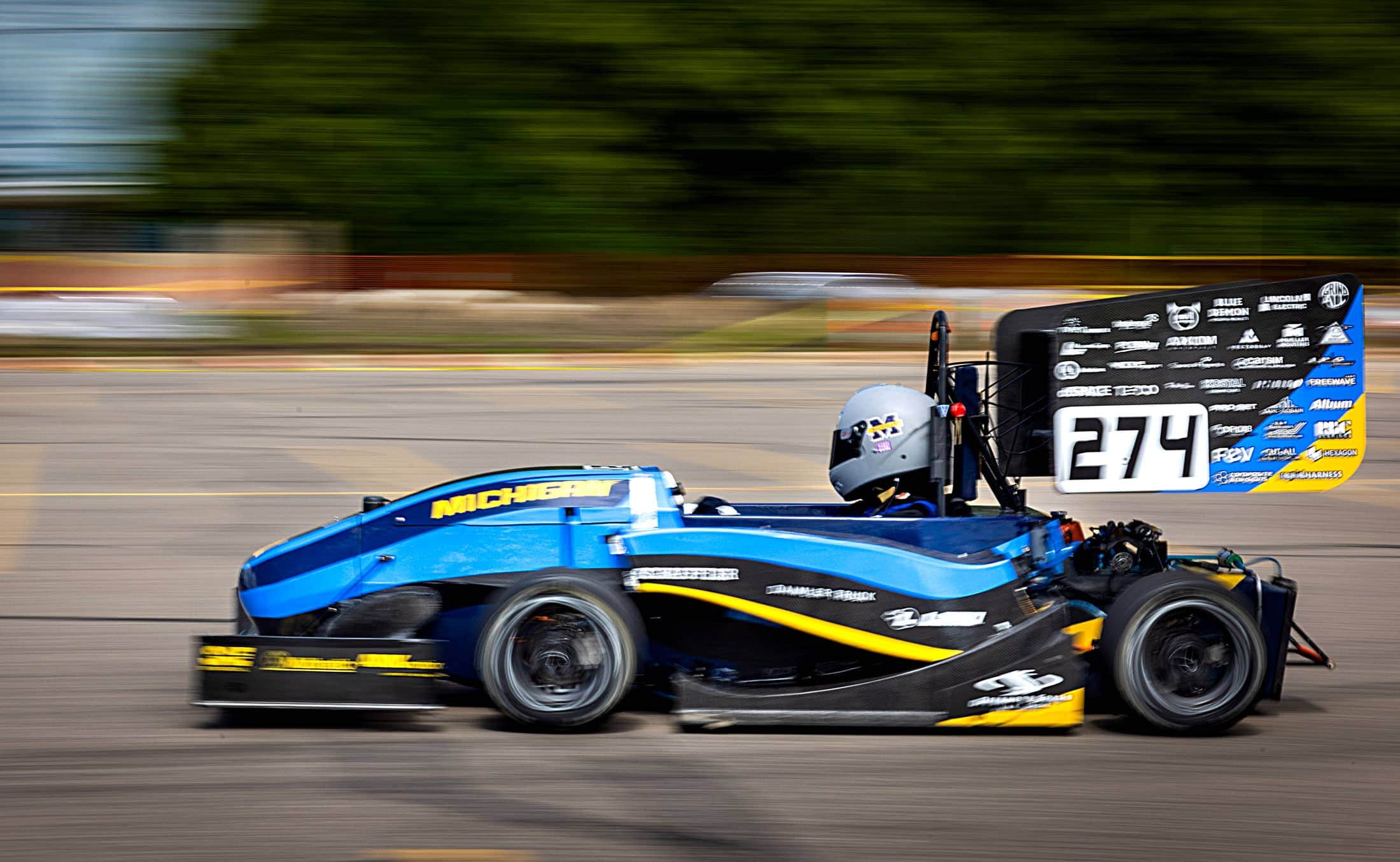 A go kart-sized race car speeds from right to left on an asphalt track.