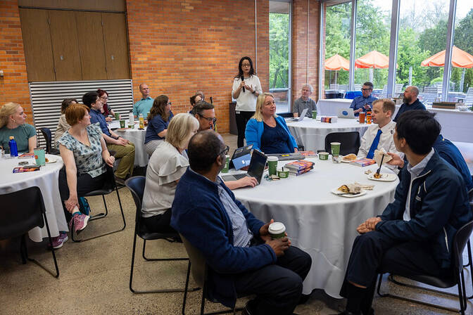 A group of people at a gathering seated around tables, with one woman standing and speaking.