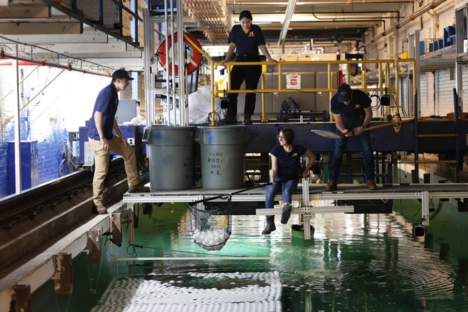 Four students are working on a platform raised above a tank of water. Two students are holding nets, which they are using to move wiffle balls from plastic barrels into the water.