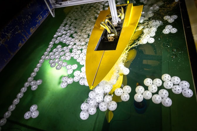 The front half of a model ship is pulled through wiffle balls floating in a long water tank. The towing carriage is above the ship and the water. The ship is connected to the carriage via a metal post.