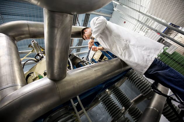 A man in a white lab coat stands next to a series of large metal tubes in a research laboratory.