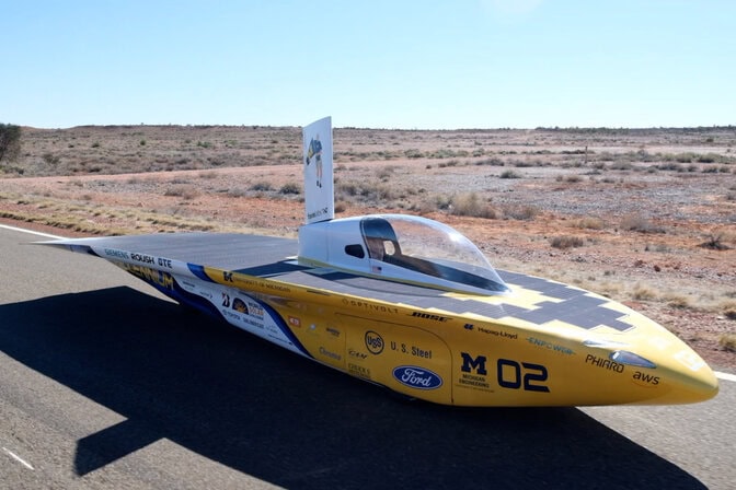 A torpedo-shaped, maize and blue car drives along a paved highway cutting through scrubby red-orange desert sand.