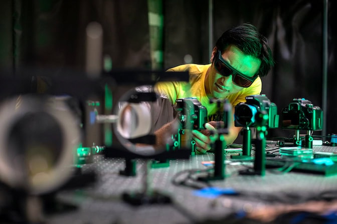 A person in safety goggles adjusts optical equipment on a lab bench under green lighting.