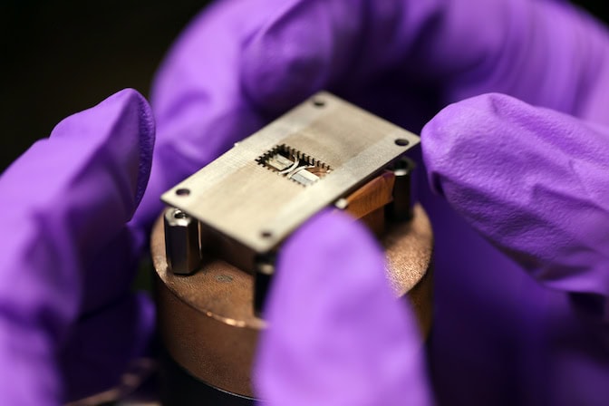 Fingers of gloved hands position a rectangular metal piece with a cutout in the center on top of a rectangular microscope standing atop a bronze cylinder.