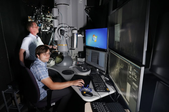 Two men work at a computer with three large screens in a dark room next to columnar microscope equipment. In the background, one man stands while looking at the microscope. In the foreground, one mat sits while monitoring a computer screen.