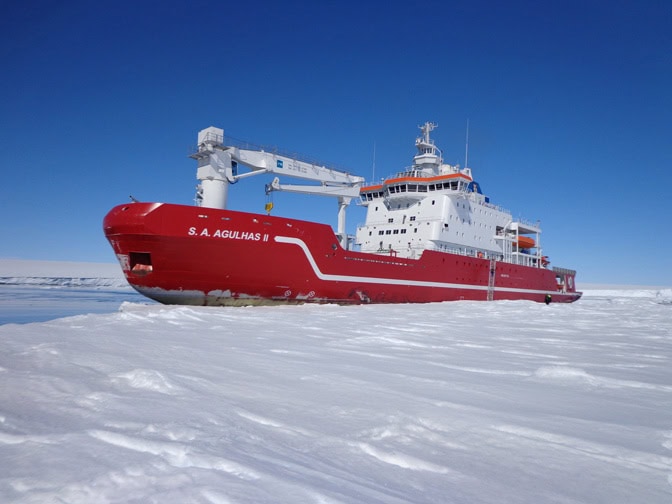 A large vessel with a red hull and white upper decks and cabins is stationed next to an ice sheet.