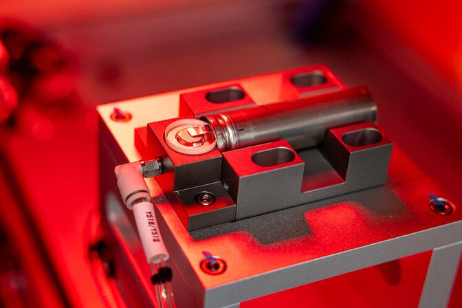 Cylindrical battery in a holder on a laser welding machine with red lighting.