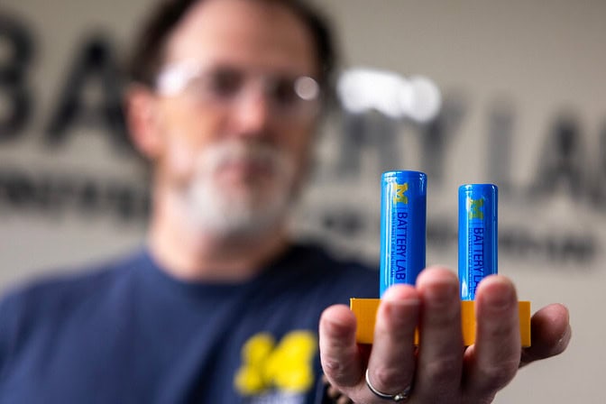A man holds two blue cylindrical batteries on an orange platform.