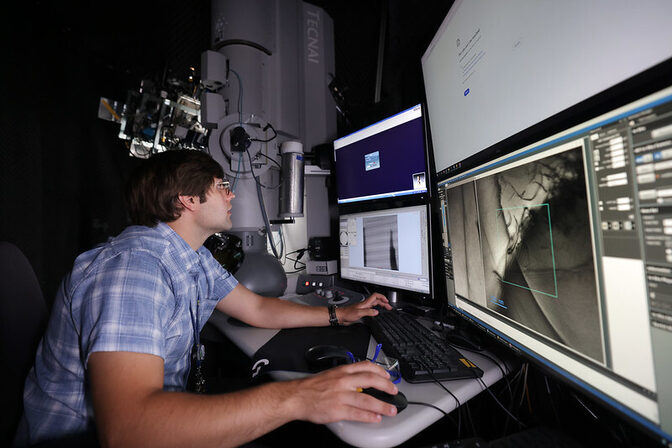 A person in a blue plaid shirt works at a workstation with three monitors displaying images and data, near advanced equipment labeled "TECNAI."