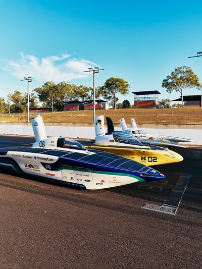 Four cars are parked in a neat row. The foremost is a blue and white torpedo with a rectangular fin protruding from the cockpit. The Michigan car, a maize-and-blue torpedo, is second in line. Its fin is similar to the first car. A flat silver car is third in line, sporting a shark-like fin on its cockpit. The fourth car is a catamaran with a fin in each of its back corners.