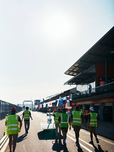 Seven members of the team's race crew walk down a long raceway. They are following their car, which is mostly knee-height save the cockpit. Flags from each of the participating countries hang from the grandstands nearby.