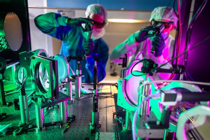 Two scientists in protective gear work with optical equipment under green and purple lights.