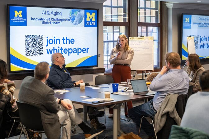 A woman stands at a conference table with people seated around and presents in front of a large display screen with a slide titled “AI & Aging: Innovations & Challenges for Global Health.”