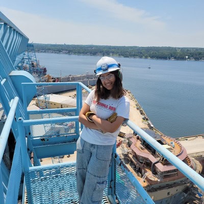 : A portrait of Archita Saraiya. She stands on a large blue gantry crane straddling a ship construction site.