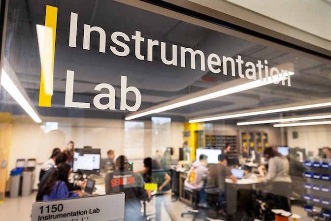 Close of a laboratory window reading, "Instrumentation Lab" with students at tables in the background out of focus.