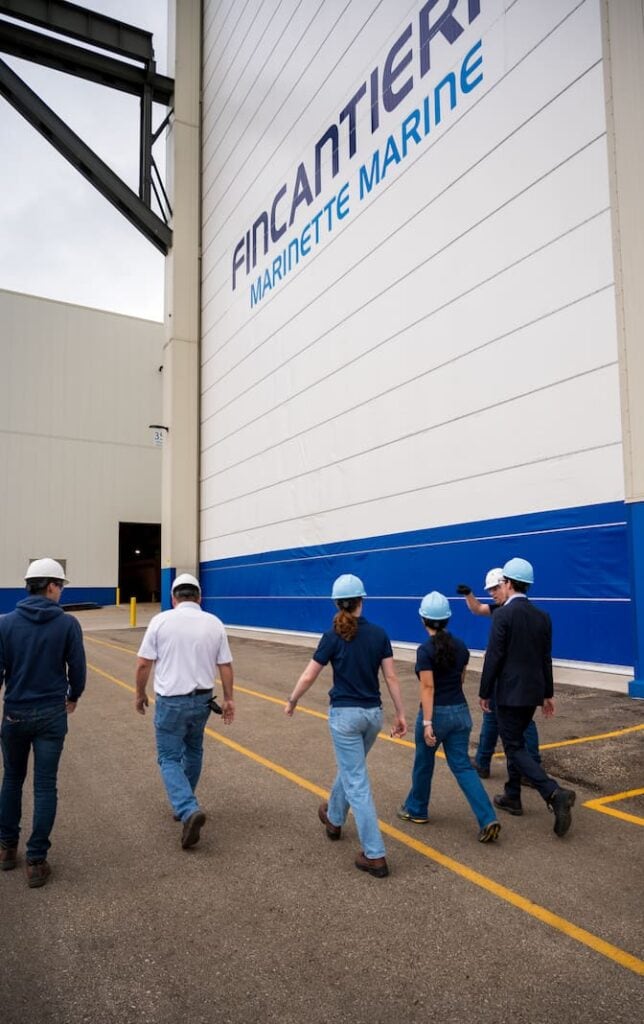 Six engineers walk past the door of a large hangar for ship construction. The shipyard name, Fincantieri Marinette Marine, is printed across the hangar door in large, blue font.