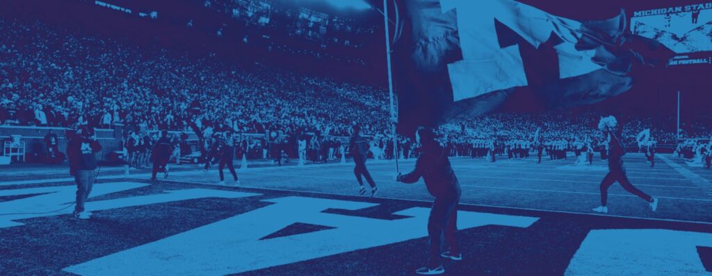 Blue overlay filter applied on a photo of a person waving a large flag with a maize University of Michigan block 'M' on it, at a crowded football game at the University of Michigan, "Big House" stadium at night.