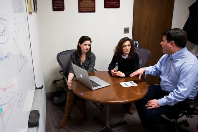Three people discussing around a table with a laptop and a whiteboard with equations in the background.