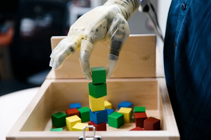 A robotic hand with a translucent covering holds a green block over a box of colorful wooden blocks.