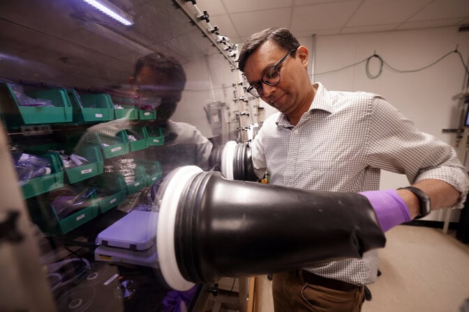 A man begins to insert a purple-gloved hand into the black glove of the box, which has trays of materials and a workspace inside.