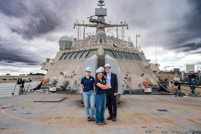 Two women and a man stand on the deck of a naval combat ship, backs facing the bridge.