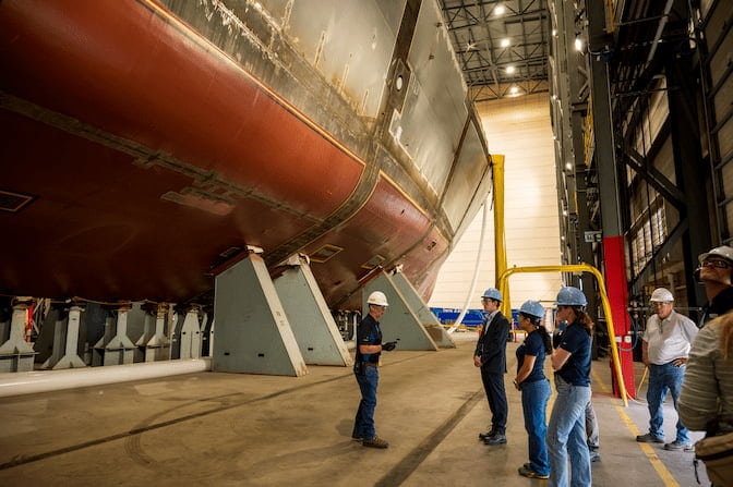 A naval vessel is supported on metal stands inside a larger hangar. The vessel's hull towers over a group of engineers standing nearby.