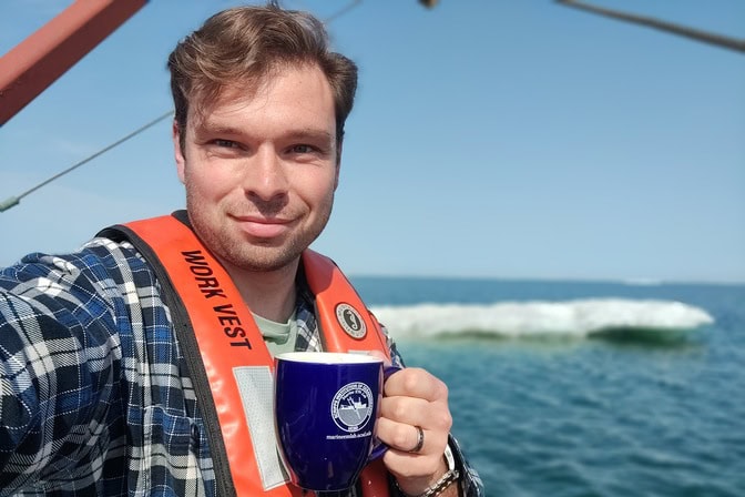 Person wearing an orange life vest on a boat, holding a blue mug, with sea and ice in the background.