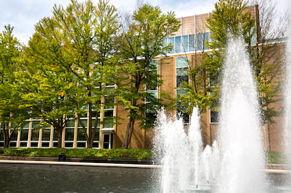 A large fountain sprays water in a rectangular pond next to a brick building with trees and people nearby.