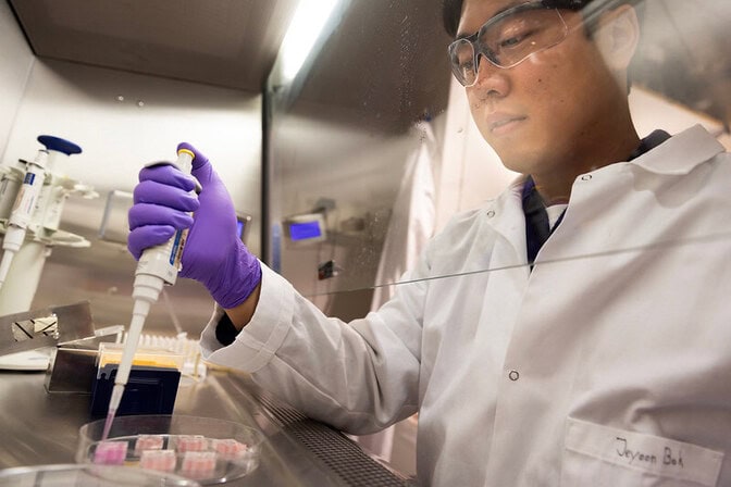 A person in a lab coat, wearing safety goggles and rubber gloves, using a pipette in a laboratory.