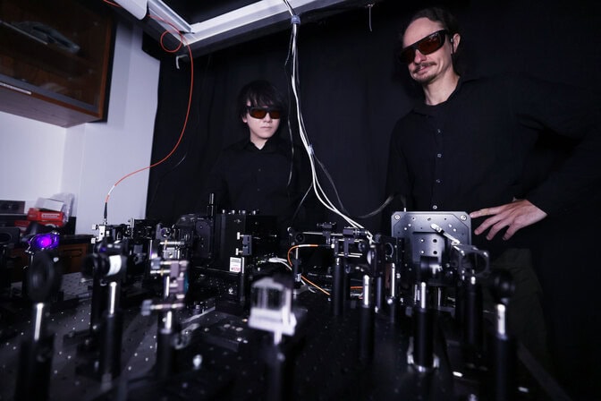 Two men in dark laser safety glasses stand behind an optical table full of silver and black optical components to guide the laser beam.