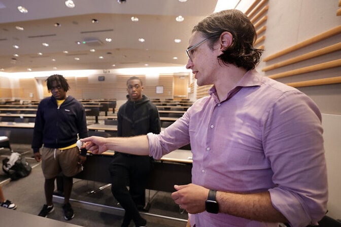 In a college lecture hall, a man in the foreground holds a ping pong ball in his hand with his arm extended while speaking. Two men in the background look on, listening.