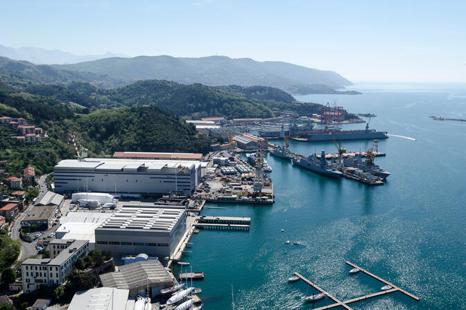 Several large buildings and hangars are situated on the shoreline. Nearby docks extend into bright, blue water. Naval vessels, yachts, and small sailboats are stationed at many of the docks.