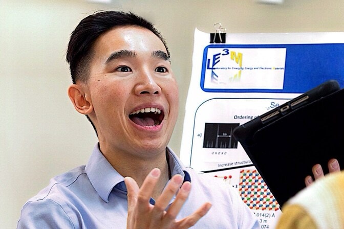 A man in a blue button down shirt wearing a red name tag speaks to an onlooker and gestures in front of an academic poster.