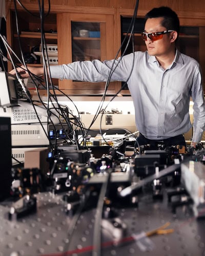 Zheshen Zhang adjusts a dial on a small screen with his arm outstretched, standing behind an optical table with a series of lenses. He wears a pair of tinted safety glasses.