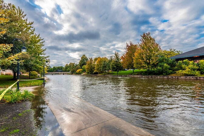 A flooded street with autumn trees bordering the street and an overcast sky. There are lamp posts partially submerged and roped off with caution tape.
