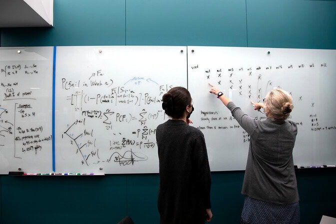 Two people discussing mathematical equations written on whiteboards.