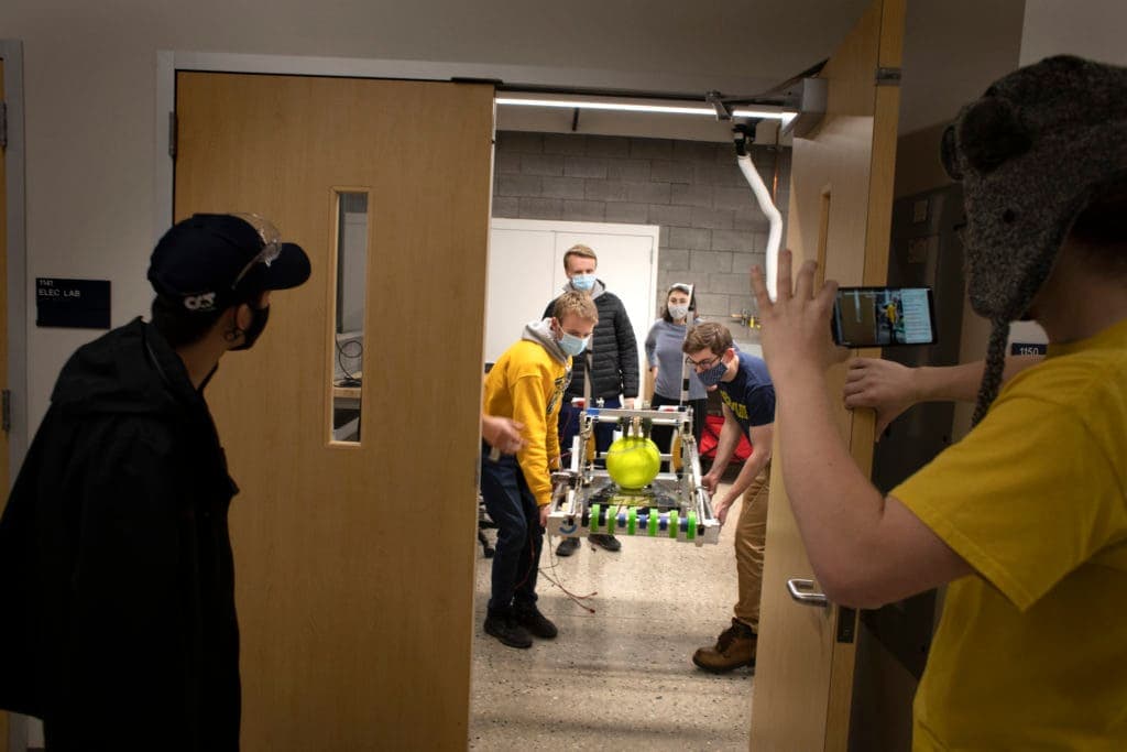 Two students carry a ball-tossing robot carefully through a propped-open door as another records the event on their phone