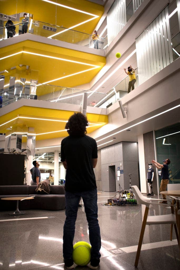 A student watches from below as another student tosses a basketball-sized tennis ball from a second-floor balcony