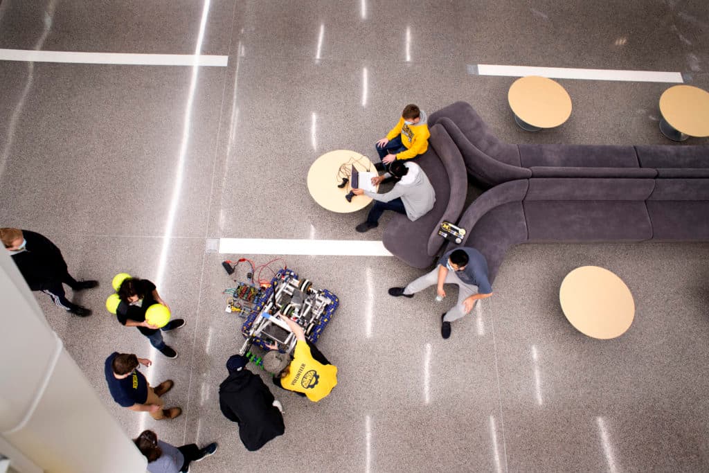 aerial view of students in an atrium lobby, some crouching around a robot, others standing and watching or seated at a small round table nearby