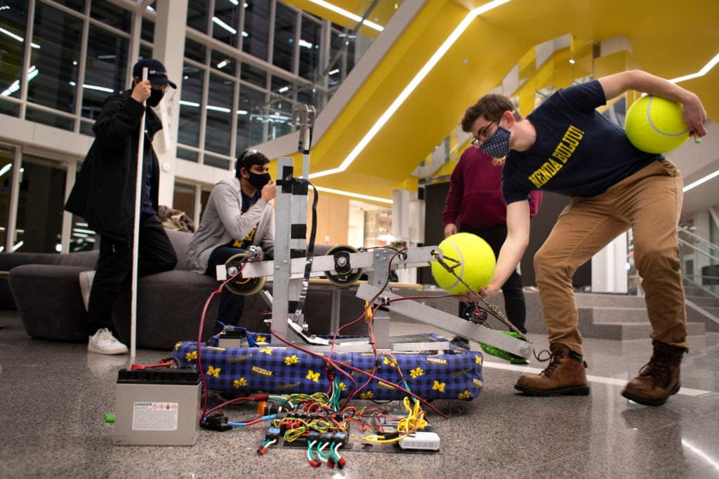 Student sets a basketball-sized tennis ball on the arm of a metal contraption with multiple levers, pulleys and wires